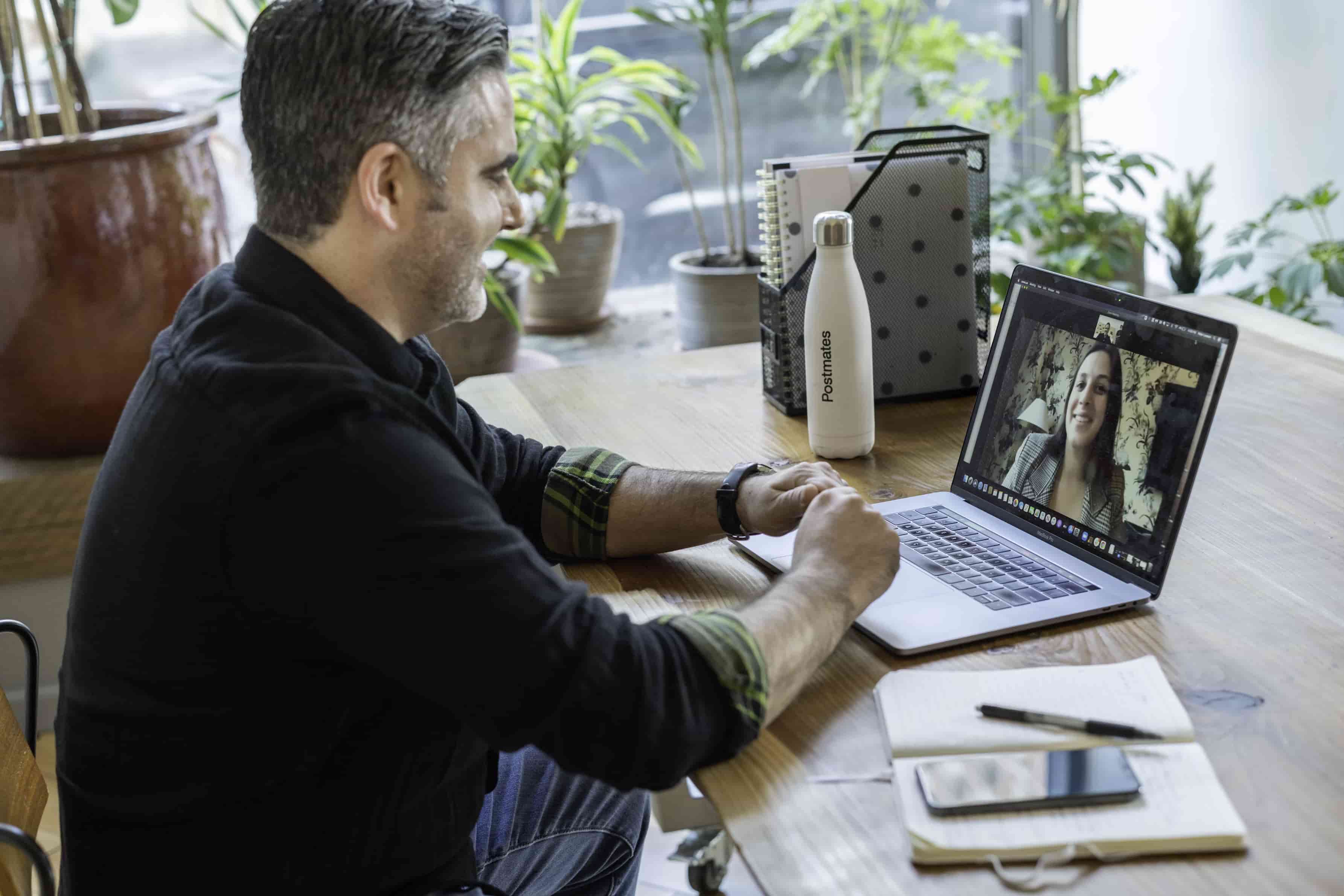 Man smiling on a video call with a woman, seated at a desk with notebook and laptop.
