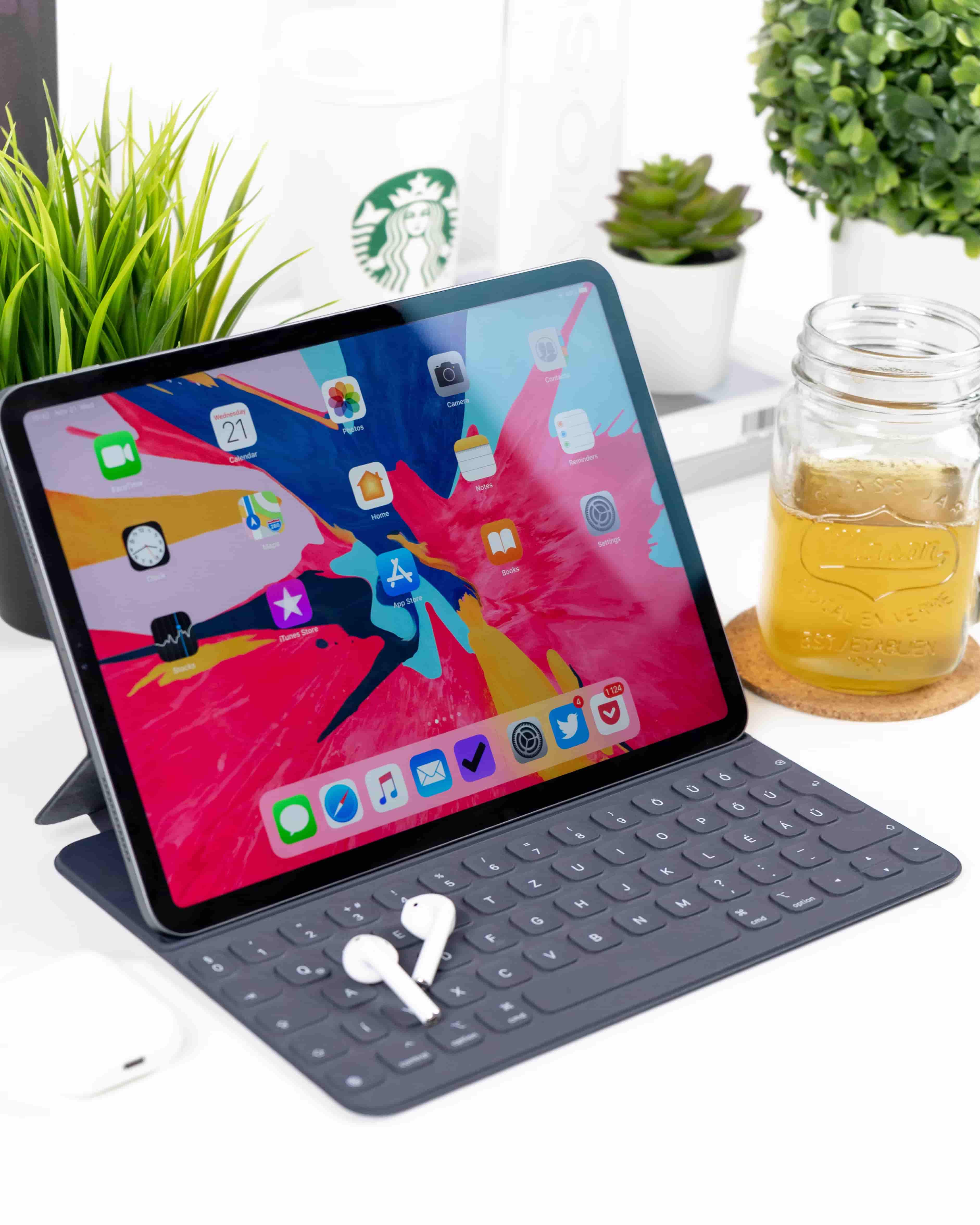 iPad with colorful home screen, Apple keyboard, AirPods, and drink on desk