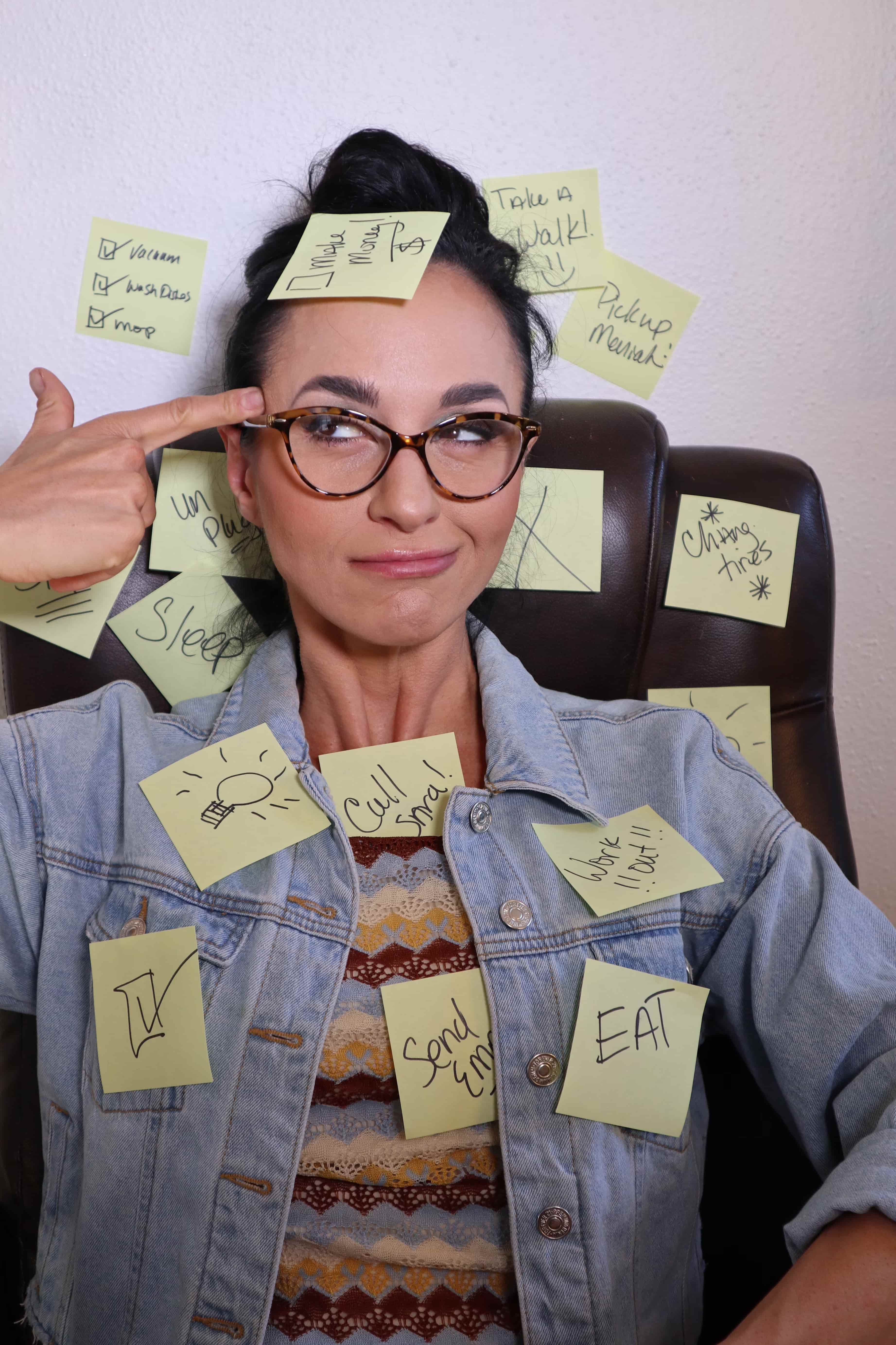 Woman wearing glasses and a denim jacket covered in sticky notes with tasks like “Work out,” “Send Email,” and “Call Sma!” She points to her temple with a sarcastic expression, illustrating overwhelm.