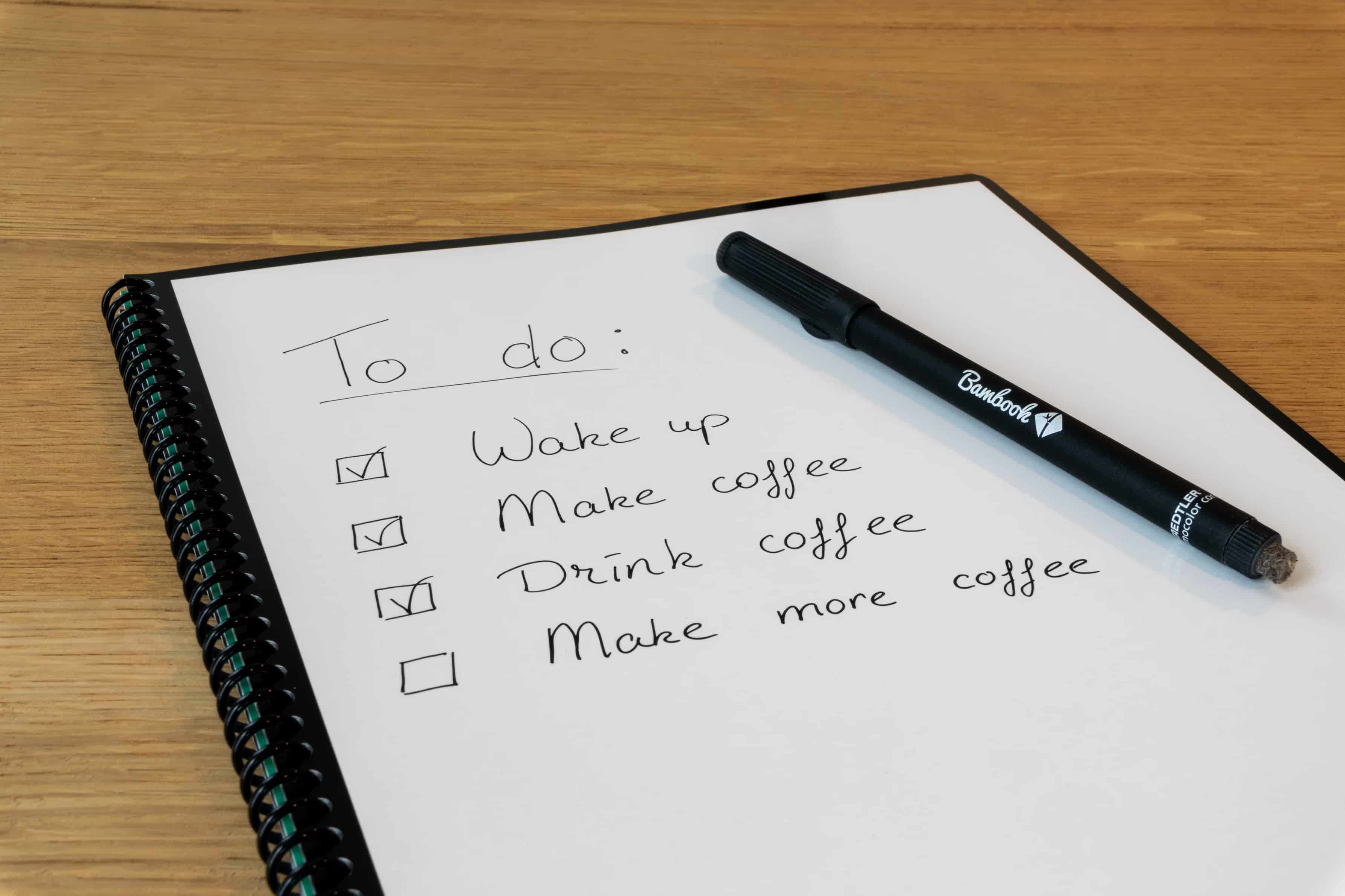 Spiral notebook with a handwritten to-do list: "Wake up, Make coffee, Drink coffee, Make more coffee." Three items are checked off. A black marker rests on the page.