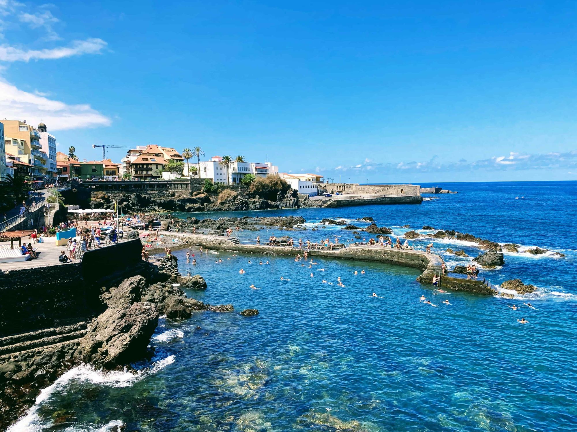 Rocky natural seawater pool in Tenerife with swimmers and a coastal town under a bright blue sky